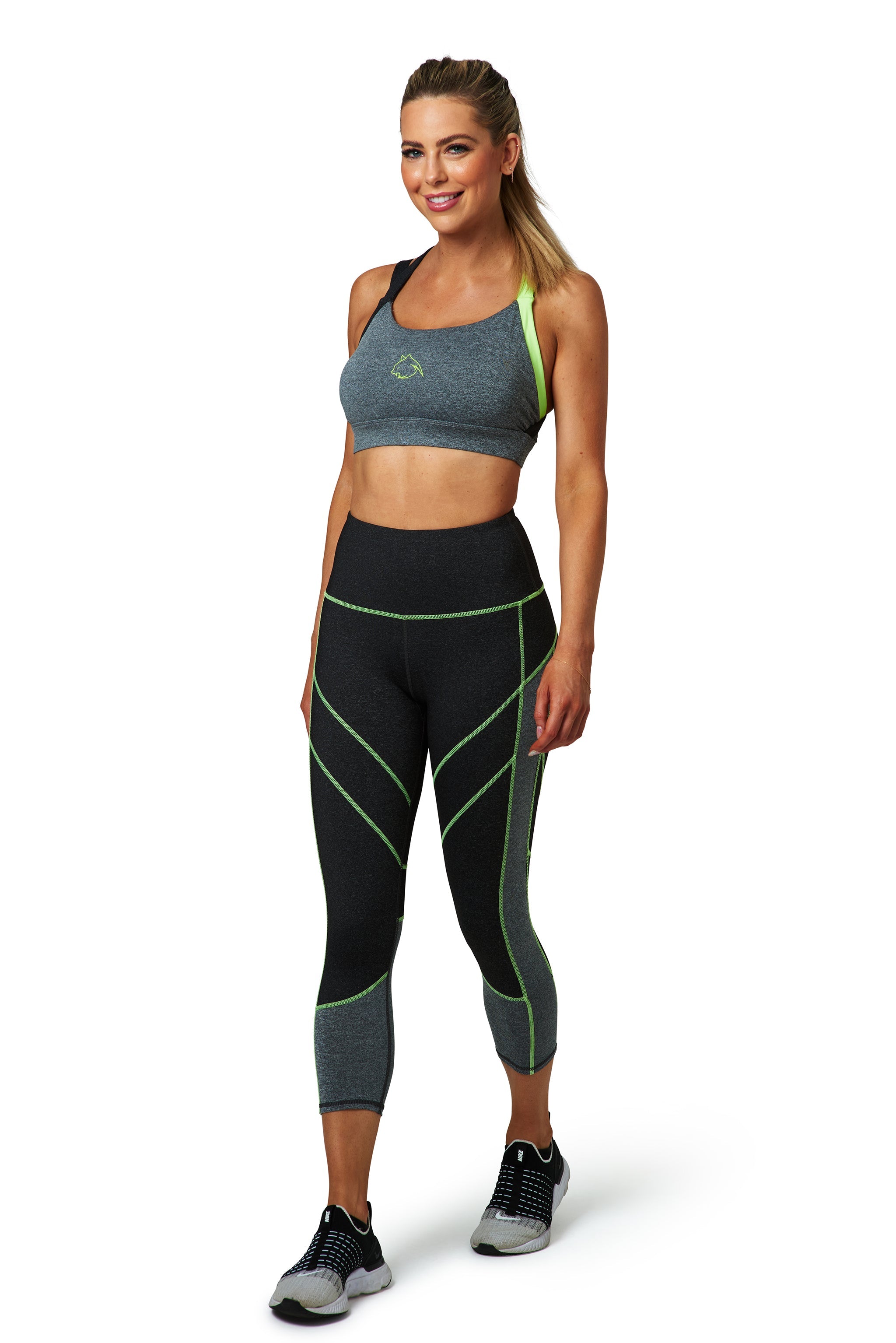 A woman wearing a BAD CAT sports bra and leggings, standing and smiling at the camera. The leggings are primarily grey with green accents and a design that provides a stay-in-place fit.